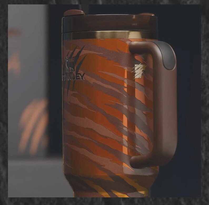 May include: A stainless steel travel mug with a tiger stripe pattern in shades of orange and brown. The mug has a brown handle, lid, and trim, and features the "Stanley" logo. The mug is designed for hot or cold beverages.