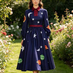 May include: A long-sleeved navy blue midi dress featuring a celestial print of white stars, crescent moons, a yellow sun, and various colourful planets. The dress has a dark red collared neckline with a button placket and a matching red waistband.