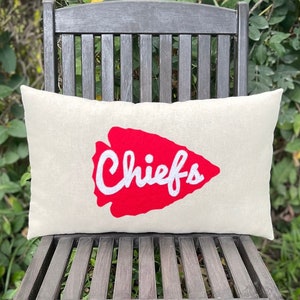 May include: A rectangular beige pillow with a red arrowhead design and the word "Chiefs" in white script. The pillow is resting on a dark wooden folding chair, set against a backdrop of green foliage and grass.