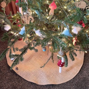 May include: A decorated Christmas tree with various ornaments, including wooden cutouts of a squirrel and raccoon, felt mountains, and mushroom-shaped decorations. The tree is surrounded by a burlap tree skirt and illuminated with string lights.