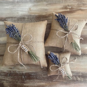 May include: Three burlap ring pillows, each adorned with a bundle of dried lavender and tied with twine. The largest pillow has a tag with the letters "K*N", while the smallest has "A+E". The pillows are arranged on a rustic wooden surface.