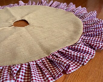 May include: A round burlap tree skirt with a ruffled edge. The skirt has a large opening in the center and is trimmed with a red and white gingham fabric. The skirt is designed for use during the holiday season.
