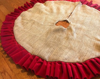May include: A round Christmas tree skirt with a burlap center and a pleated red velvet border. The skirt has a hole in the center for the tree trunk. The skirt is designed to sit on the floor around the base of a Christmas tree.