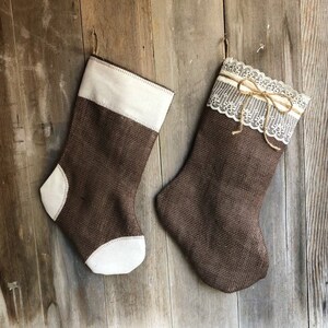 May include: Two brown burlap Christmas stockings with off-white details. One has a white cuff, the other a white lace cuff with a twine bow. Both are suspended against a rustic wooden backdrop.