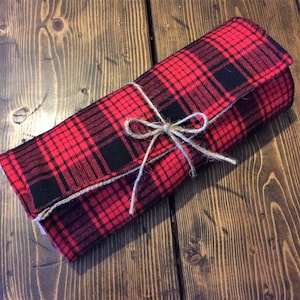 May include: A red and black plaid fabric roll tied with twine. The fabric has a classic tartan pattern. The roll is closed with a natural twine bow. The item is on a wooden surface.