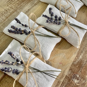 Puede incluir: Varios peque&ntilde;os saquitos rectangulares de lino blanco est&aacute;n dispuestos sobre una superficie de madera. Cada saquito est&aacute; atado con hilo de bramante y adornado con un manojo de lavanda seca. La lavanda tiene un tono p&uacute;rpura intenso y el hilo es de color marr&oacute;n natural.