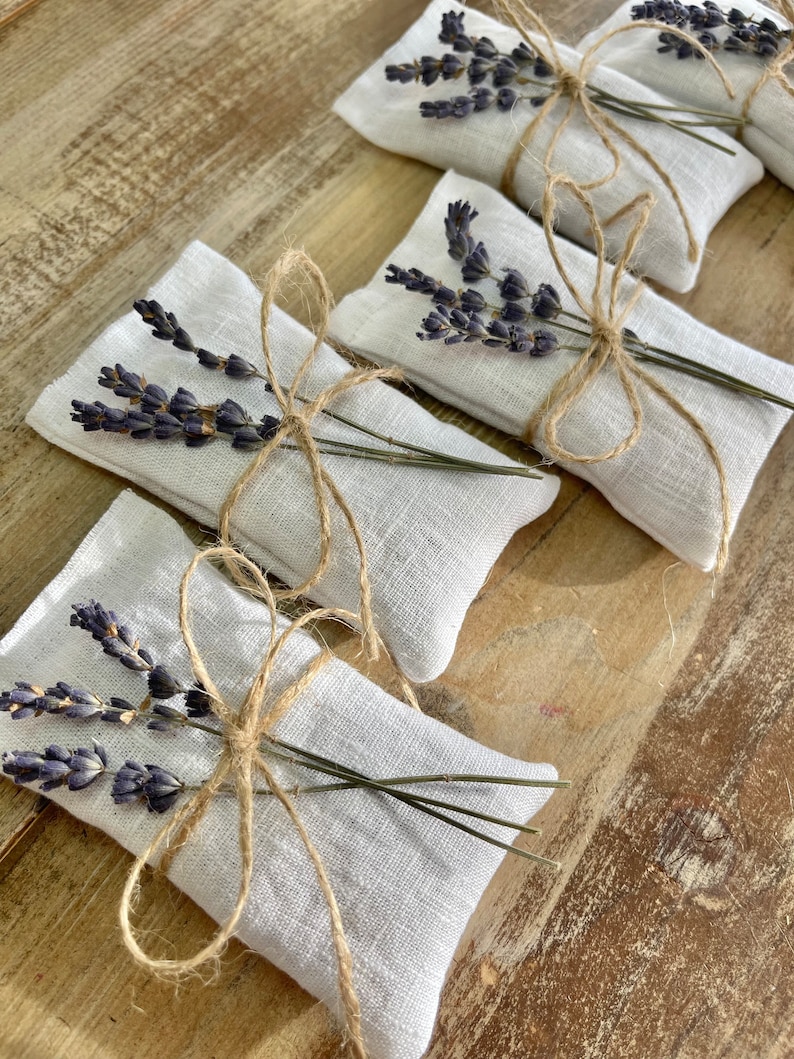 Puede incluir: Varios peque&ntilde;os saquitos rectangulares de lino blanco est&aacute;n dispuestos sobre una superficie de madera. Cada saquito est&aacute; atado con hilo de bramante y adornado con un manojo de lavanda seca. La lavanda tiene un tono p&uacute;rpura intenso y el hilo es de color marr&oacute;n natural.