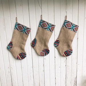May include: Three burlap Christmas stockings with Southwestern-style patterned tops and toe accents. The stockings are tan with a brown, turquoise, red, and white geometric design. Each stocking hangs from a twine loop.