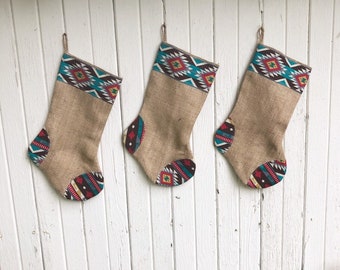 May include: Three burlap Christmas stockings with Southwestern-style patterned tops and toe accents. The stockings are tan with a brown, turquoise, red, and white geometric design. Each stocking hangs from a twine loop.