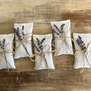 Puede incluir: Cinco peque&ntilde;os saquitos rectangulares de lino blanco, cada uno atado con hilo y adornado con una ramita de lavanda seca. Los saquitos est&aacute;n dispuestos sobre una superficie de madera r&uacute;stica de color marr&oacute;n claro.