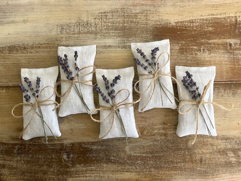 Puede incluir: Cinco peque&ntilde;os saquitos rectangulares de lino blanco, cada uno atado con hilo y adornado con una ramita de lavanda seca. Los saquitos est&aacute;n dispuestos sobre una superficie de madera r&uacute;stica de color marr&oacute;n claro.