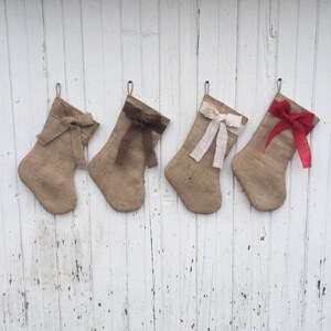 May include: Four burlap Christmas stockings with different coloured bows hanging on a white wooden wall. The bows are in shades of brown, white, and red. Each stocking has a loop for hanging.
