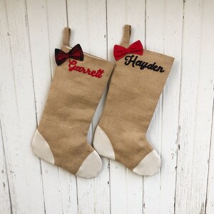 May include: Two burlap Christmas stockings with white toes and hanging loops. One stocking has a red and black plaid bow and the name "Garrett" in red script. The other has a red bow and the name "Hayden" in black script.