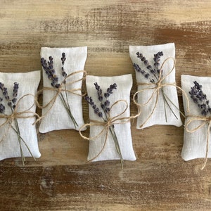 Puede incluir: Cinco saquitos de lino blanco rellenos de lavanda seca, cada uno atado con hilo de bramante y una ramita de lavanda. Los saquitos est&aacute;n dispuestos sobre una superficie de madera r&uacute;stica de color marr&oacute;n claro.