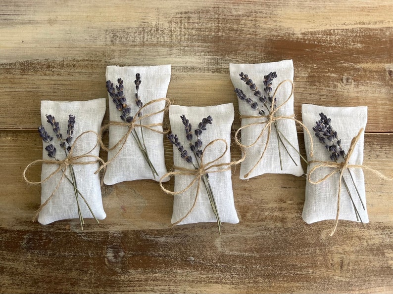 Puede incluir: Cinco saquitos de lino blanco rellenos de lavanda seca, cada uno atado con hilo de bramante y una ramita de lavanda. Los saquitos est&aacute;n dispuestos sobre una superficie de madera r&uacute;stica de color marr&oacute;n claro.