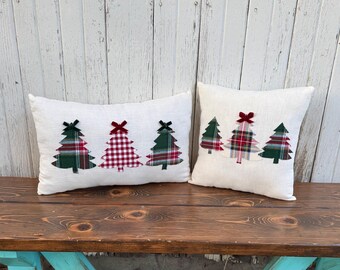 May include: Two decorative Christmas pillows with white linen fabric. Each pillow features appliqued Christmas trees in green and red plaid patterns, with red bow accents. The pillows are displayed on a wooden bench.