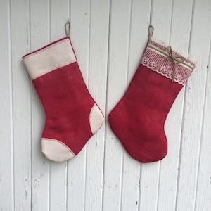 May include: Two red Christmas stockings with off-white accents hang against a white wooden background. One stocking has a solid off-white cuff and toe, while the other has a lace and ribbon cuff.