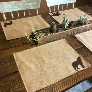 Puede incluir: Juegos de manteles individuales rectangulares de arpillera con siluetas de animales marrones, como un lobo, un oso y un ciervo. Una jardinera de madera con suculentas y cactus.