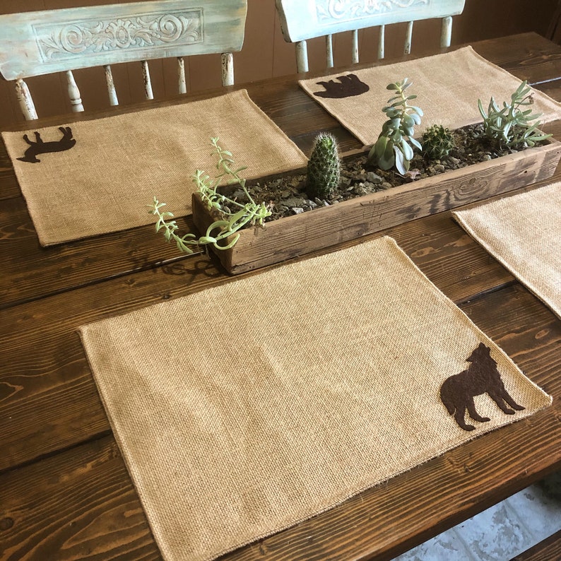 Puede incluir: Juegos de manteles individuales rectangulares de arpillera con siluetas de animales marrones, como un lobo, un oso y un ciervo. Una jardinera de madera con suculentas y cactus.
