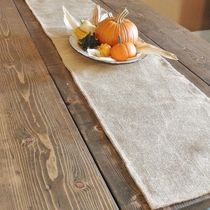 May include: A burlap table runner on a dark wood table. A white plate holds an arrangement of pumpkins and gourds in shades of orange and yellow. The table has a natural wood grain.