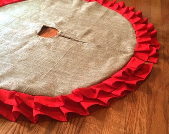 May include: A round burlap Christmas tree skirt with a vibrant red ruffled edge. The skirt has a hole in the center for the tree trunk and is displayed on a hardwood floor. The neutral tones of the burlap contrast with the bright red.