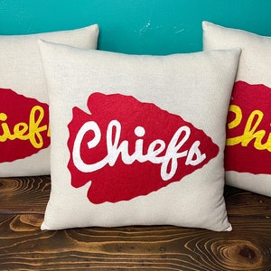 May include: Three square throw pillows with a cream-colored background. Each pillow features a red arrowhead design with the word "Chiefs" in white script. The pillows are arranged on a dark wooden surface.