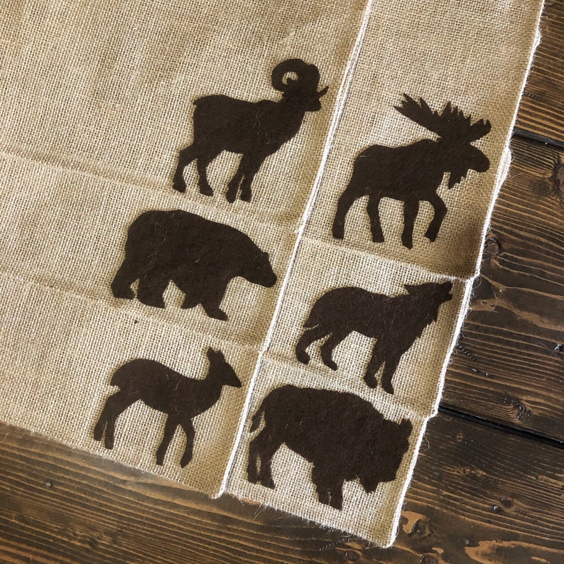 Puede incluir: Un camino de mesa de arpillera con siluetas de animales marr&oacute;n oscuro. El camino de mesa presenta un carnero, un alce, un oso, un lobo, un ciervo y un bisonte. El camino de mesa est&aacute; sobre una superficie de madera oscura.