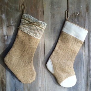 May include: Two burlap Christmas stockings hanging against a weathered wooden background. One stocking features a lace trim and a twine bow, while the other has a white cuff and toe. Both have a loop for hanging.