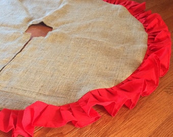 May include: A round burlap Christmas tree skirt with a bright red ruffled edge. The skirt has a split opening and a center hole for the tree trunk. The natural tan color of the burlap contrasts with the vibrant red.