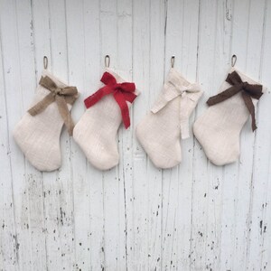 May include: Four cream Christmas stockings with burlap bows in brown, red, white, and dark brown. The stockings hang from metal hooks against a weathered white wooden background.