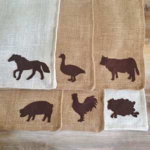 May include: A collection of burlap placemats with dark brown animal silhouettes. The placemats feature a horse, goose, cow, pig, rooster, and sheep. The placemats are a mix of natural burlap and off-white burlap.