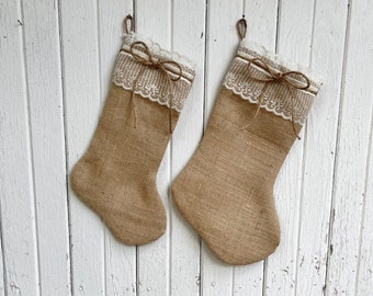May include: Two burlap Christmas stockings with white lace trim and twine bows hanging against a white wooden background. The stockings have a rustic, farmhouse style, perfect for holiday decor.