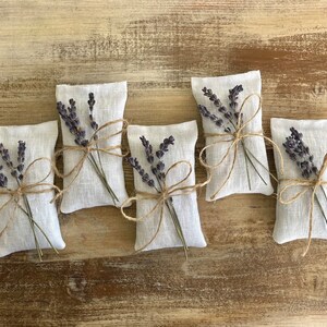 Puede incluir: Cinco peque&ntilde;os saquitos rectangulares de lino blanco rellenos de lavanda seca. Cada saquito est&aacute; atado con hilo de bramante y adornado con una ramita de lavanda. El conjunto est&aacute; sobre una superficie de madera r&uacute;stica marr&oacute;n claro.
