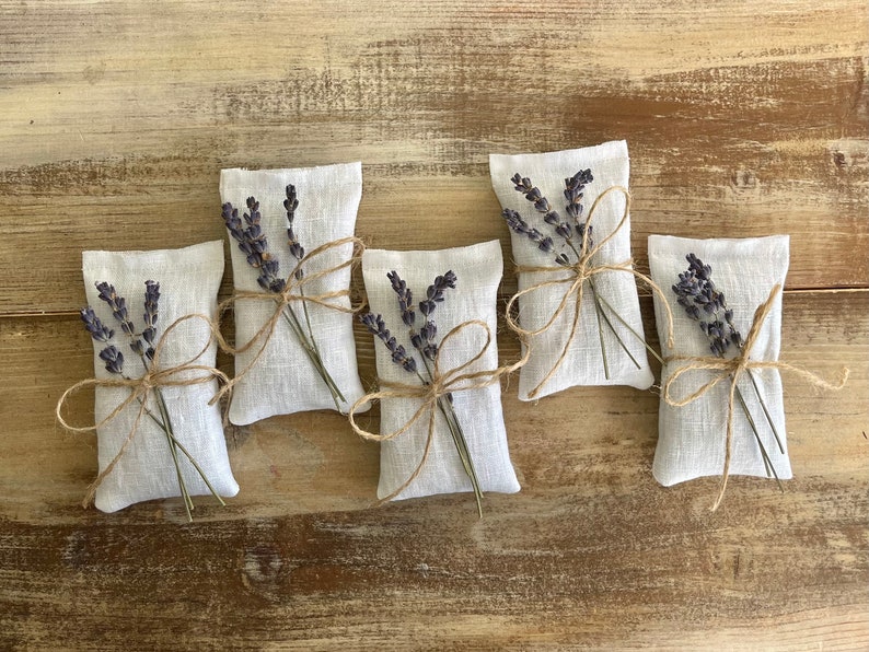Puede incluir: Cinco peque&ntilde;os saquitos rectangulares de lino blanco rellenos de lavanda seca. Cada saquito est&aacute; atado con hilo de bramante y adornado con una ramita de lavanda. El conjunto est&aacute; sobre una superficie de madera r&uacute;stica marr&oacute;n claro.