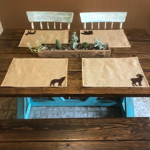 Puede incluir: Una mesa de comedor r&uacute;stica con cuatro manteles individuales de arpillera, cada uno con una silueta de animal diferente: un ciervo, un oso, un lobo aullando y un carnero. Una jardinera de madera con suculentas y cactus se encuentra en el centro. Dos sillas azul claro est&aacute;n en el fondo.