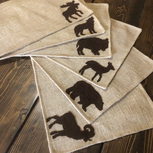 Puede incluir: Juego de seis manteles individuales de arpillera con siluetas de animales de fieltro marr&oacute;n: alce, lobo, bisonte, ciervo, oso y carnero. Los manteles individuales est&aacute;n dispuestos en forma de abanico, mostrando la textura r&uacute;stica.