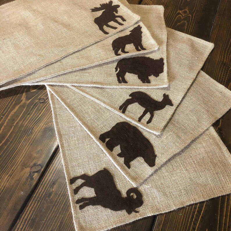 Puede incluir: Juego de seis manteles individuales de arpillera con siluetas de animales de fieltro marr&oacute;n: alce, lobo, bisonte, ciervo, oso y carnero. Los manteles individuales est&aacute;n dispuestos en forma de abanico, mostrando la textura r&uacute;stica.
