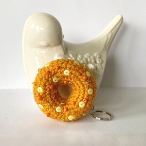 May include: A crocheted donut keyring in shades of yellow and orange, decorated with sequins and beads. The donut is round with a hole in the middle and attached to a silver keyring. A white ceramic bird is in the background.