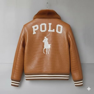 Può includere: Giacca in pelle marrone con colletto in ecopelliccia. La parte posteriore presenta la scritta "POLO" in lettere bianche a blocchi sopra un logo di giocatore di polo ricamato bianco. La giacca ha una texture a coccodrillo e polsini e orlo a righe.
