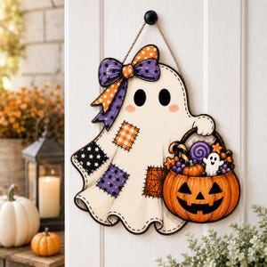 May include: A decorative wall hanging shaped like a white ghost with black eyes and pink cheeks. The ghost wears a purple and orange polka dot bow and features patches with stars, plaid, and spiderweb patterns, while holding a jack-o'-lantern bucket.