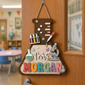 Könnte beinhalten: Ein dunkelbraunes Holzschild in Form eines Laborkolbens mit dem Text "mrs" und "MORGAN" in bunten Buchstaben. Es enthält 3D-Ausschnitte von Reagenzgläsern, einem Mikroskop, einer Pipette, einer Lupe und kleinen Sternen.