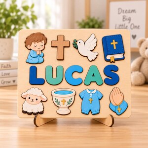 May include: A rectangular wooden puzzle board featuring the name LUCAS in large blue and teal letters. Surrounding the name are wooden cutouts of a praying child, a brown cross, a white dove, a blue Bible, a lamb, a baptismal font, a blue outfit, and hands.