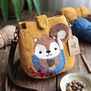 May include: A mustard yellow punch needle crossbody bag featuring a textured squirrel illustration holding an acorn. The bag has a brown leather strap, a flap closure, and a small tag with an acorn graphic. The squirrel is depicted in brown, white, and blue tones with red floral accents at the bottom.