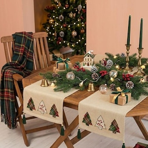 May include: A beige fabric table runner featuring a repeating pattern of embroidered Christmas trees in red and green plaid fabric, finished with green tassels at the ends. The runner is decorated with pine branches, ornaments, and candles.