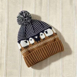 May include: A knit beanie featuring a brown ribbed cuff and a dark blue upper section with a white sheep pattern. The hat is topped with a matching dark blue and white pom-pom, showcasing a detailed jacquard knit construction throughout.