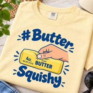 May include: A pale yellow t-shirt featuring a graphic of a hand squeezing a stick of butter. The text reads #Butter and Squishy in blue, with the butter label stating 4oz. NET WT. (113 G) SALTED BUTTER. The shirt has a Comfort Colors brand tag.