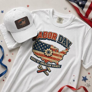 May include: A white t-shirt and a matching white baseball cap. The shirt features a graphic of the United States map with a flag pattern, a gear icon, and the text LABOR DAY BUILT BY AMERICAN HANDS. The cap has a rectangular patch with the same design.