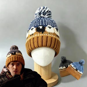 May include: A knit beanie featuring a pattern of white and black sheep against a blue background, with a solid mustard-yellow ribbed cuff and a blue and white pom-pom on top. The design includes a white geometric border above the sheep.