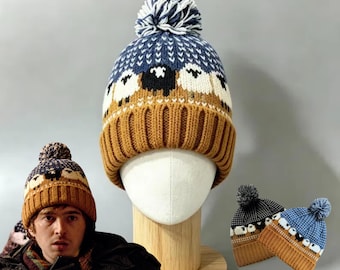 Puede incluir: Un gorro de punto con un patrón de ovejas blancas y negras sobre un fondo azul, con un puño acanalado de color amarillo mostaza sólido y un pompón azul y blanco en la parte superior. El diseño incluye un borde geométrico blanco sobre las ovejas.