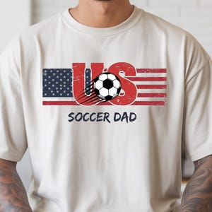 May include: A beige short-sleeved t-shirt featuring a graphic design with the text US, where the U is styled like the American flag and the S is red. A black and white soccer ball with motion lines sits in the center, with SOCCER DAD below.